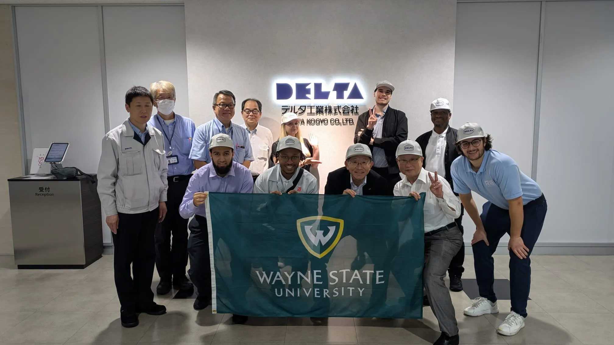 Wayne State students and DELTA staff posed together holding a Wayne State flag in front of the DELTA company sign in the lobby.