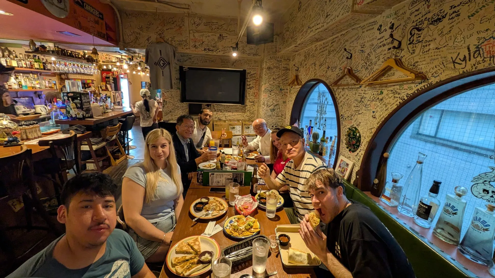 The group crowded around a table in a small restaurant with graffiti-covered walls and round porthole windows, plates and drinks in front of them.