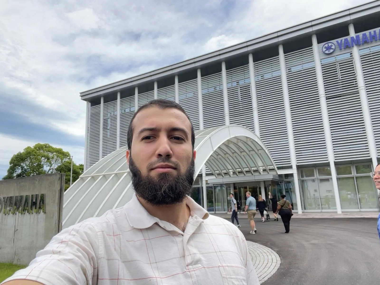 Gotibeh outside the glass and steel entrance of the Yamaha factory under a cloudy sky.