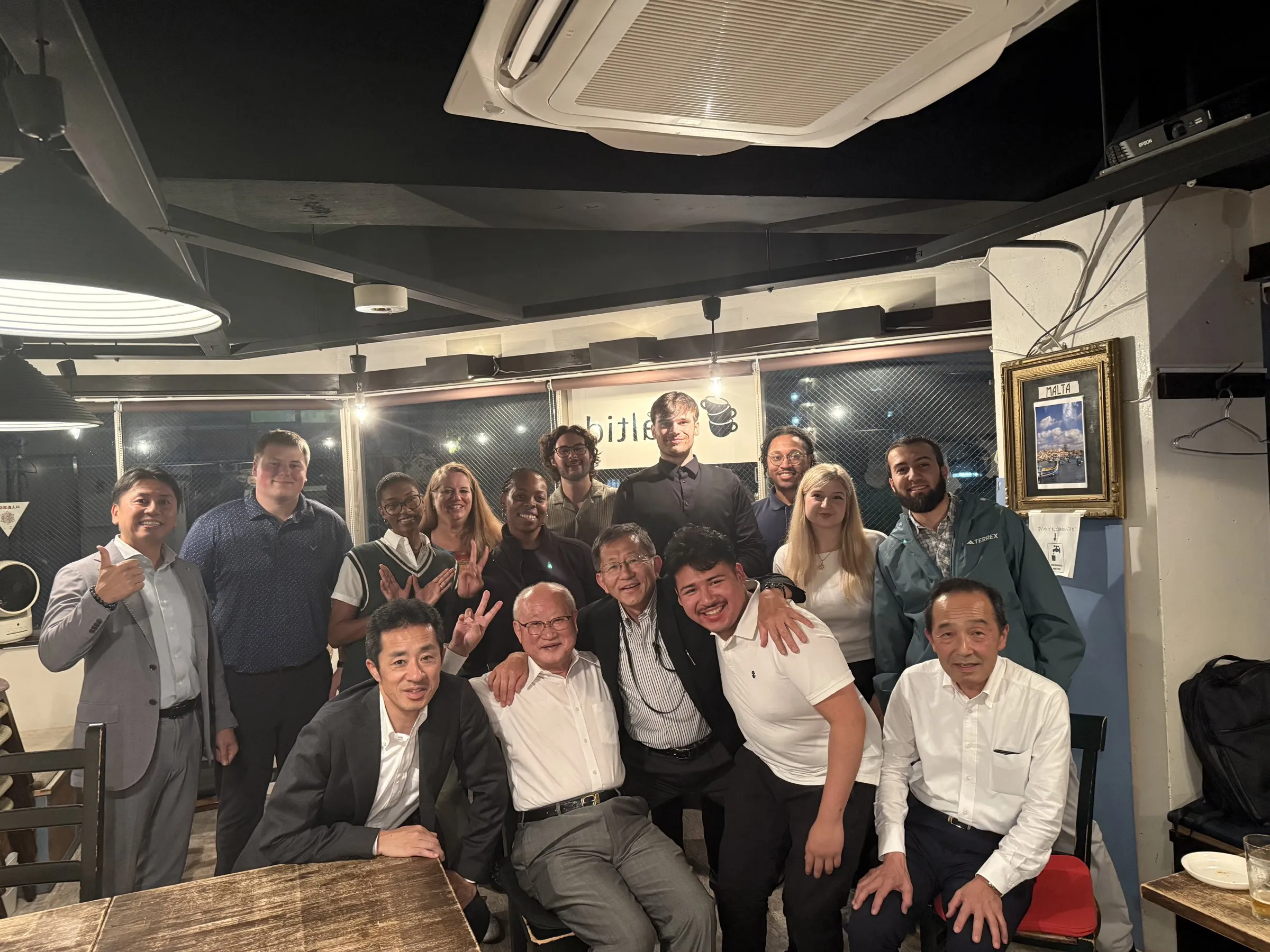 A large group of students and Japanese hosts packed together for a photo at the end of a dinner in a small restaurant.