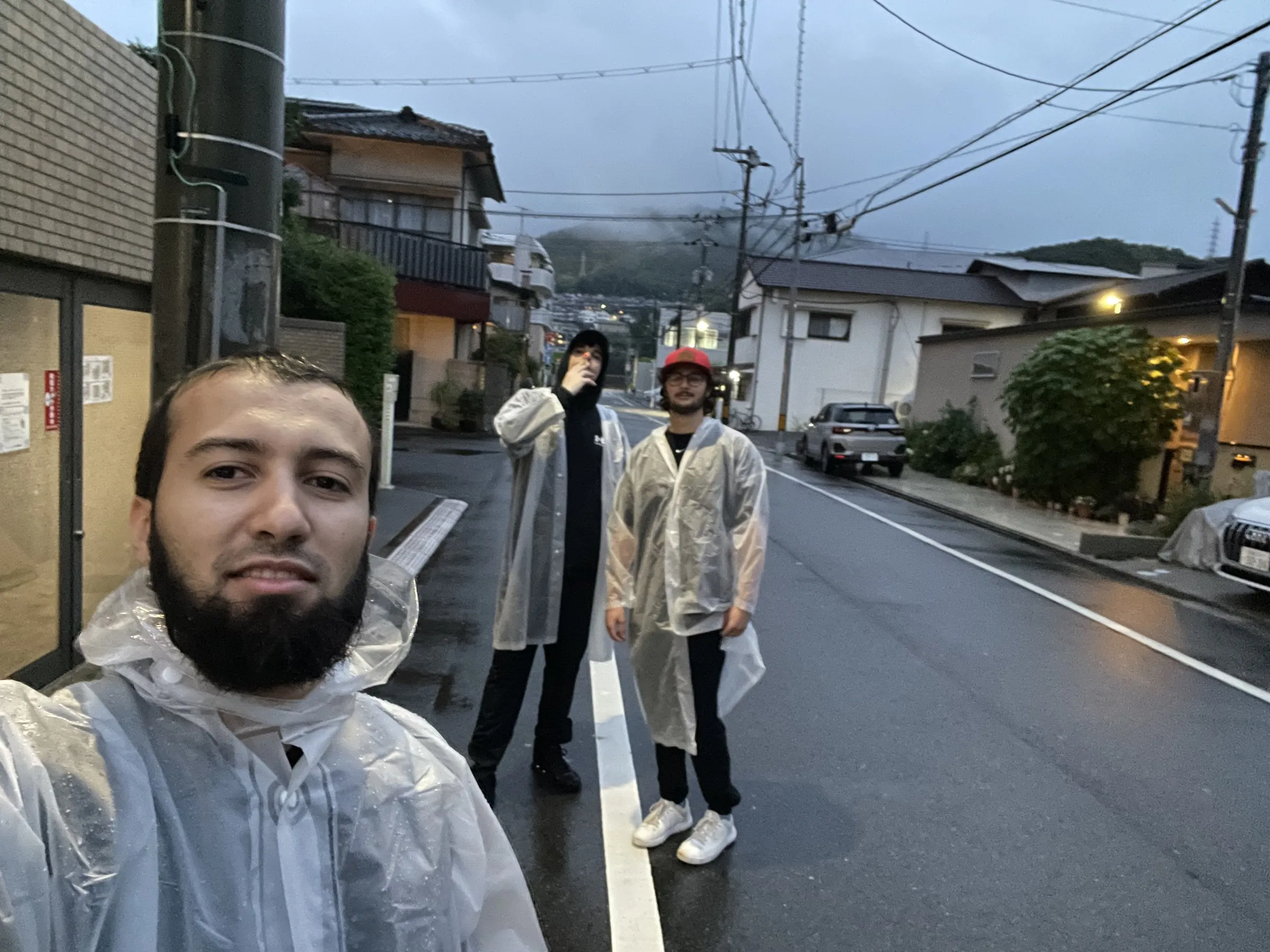 Three classmates in clear plastic rain ponchos standing on a wet residential street in Hiroshima at dusk.