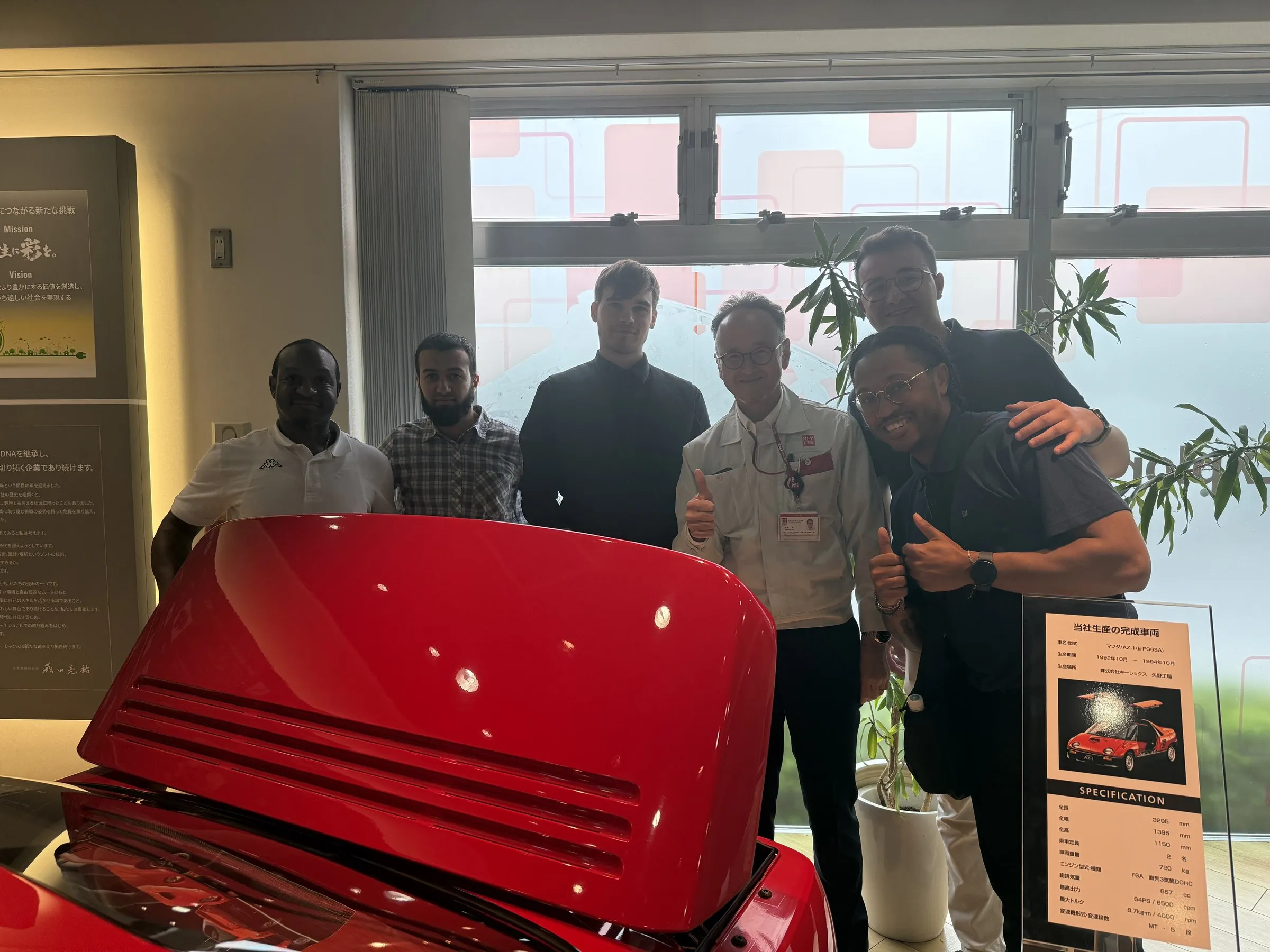Students and a Keylex host standing behind a red Mazda AZ-1 with its gullwing door raised, beside a specification placard.