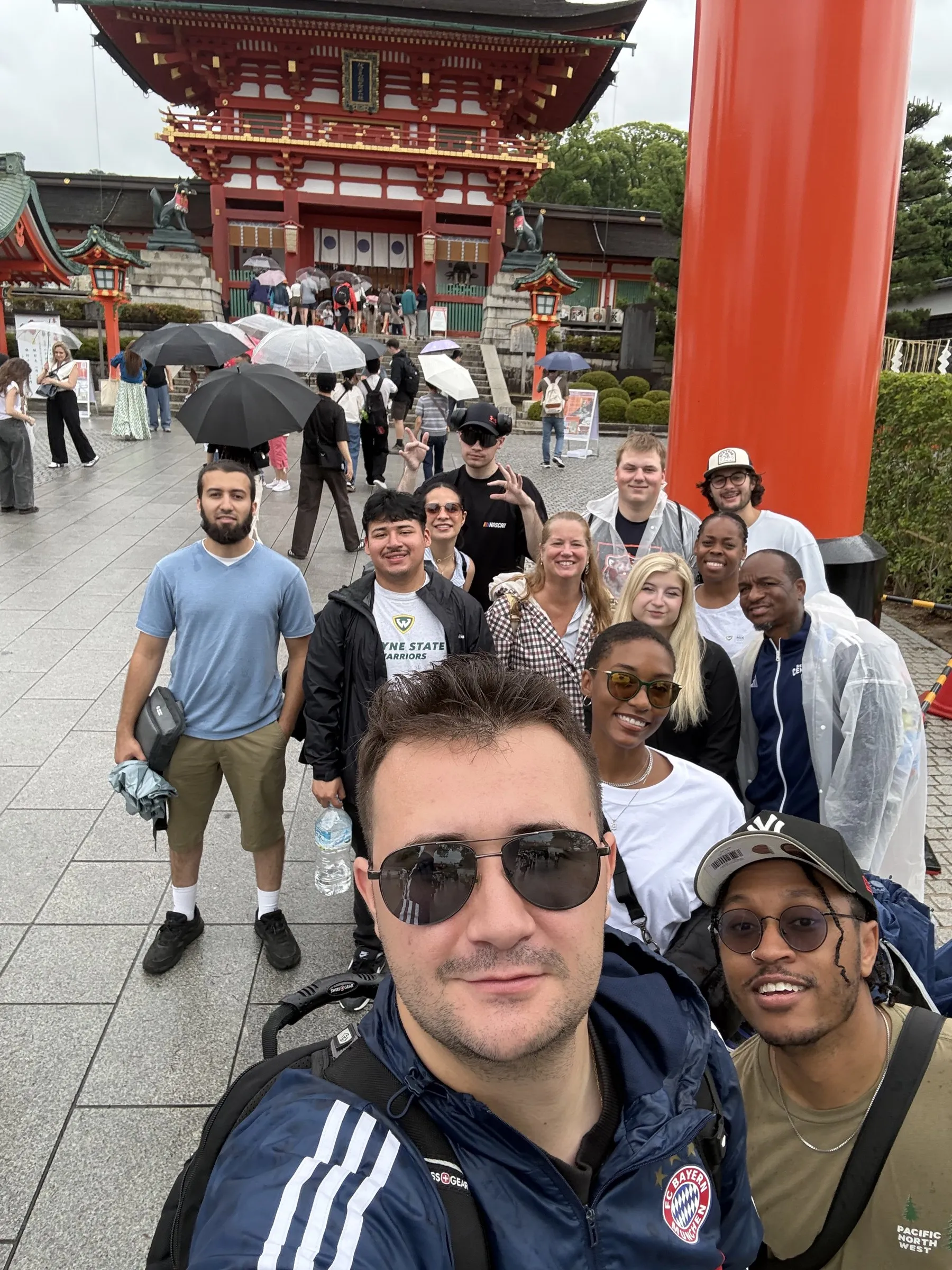 A group selfie of the Wayne State students in front of the main gate at Fushimi Inari shrine in Kyoto, with umbrellas up in the rain.