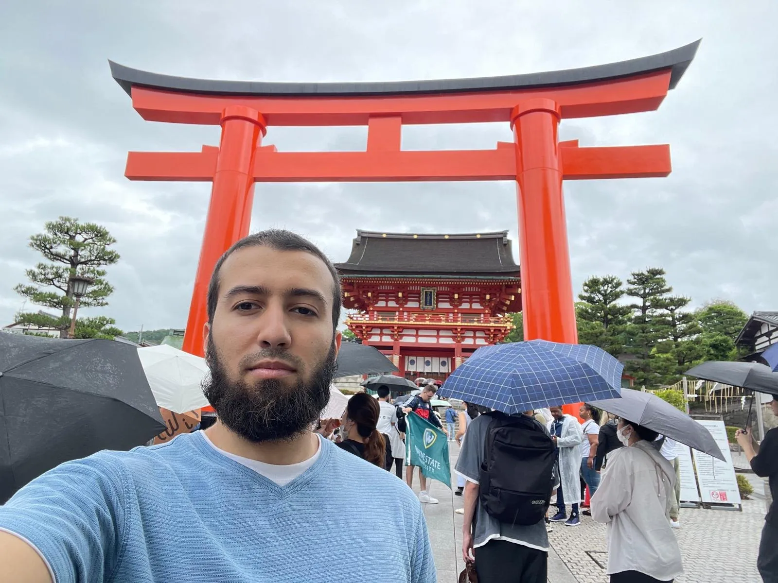 Gotibeh in a blue polo beside a tall vermilion torii pillar at Fushimi Inari, the shrine buildings behind him.