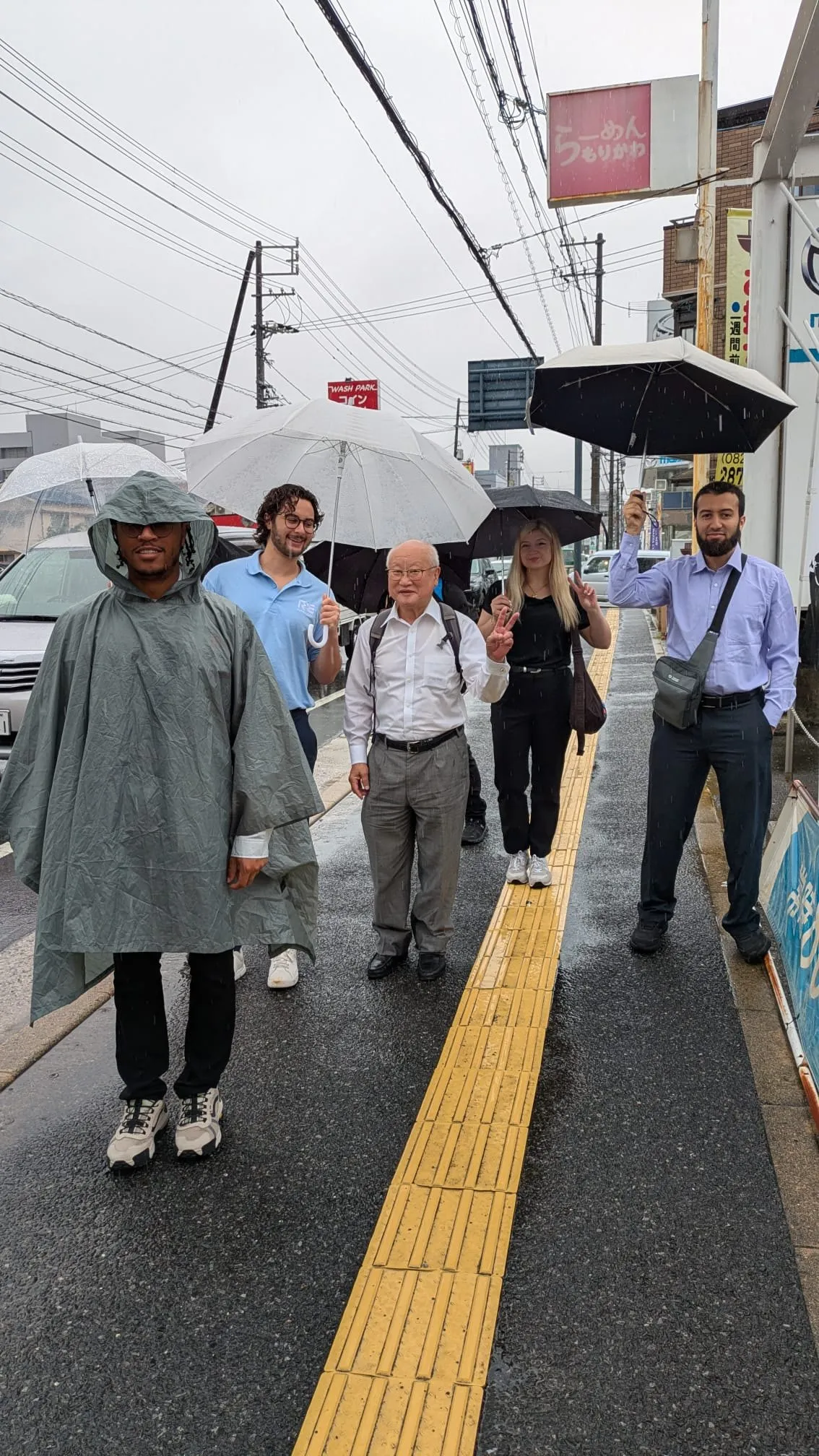 The group walking in ponchos and under umbrellas along a wet street in Hiroshima, following the yellow tactile paving.