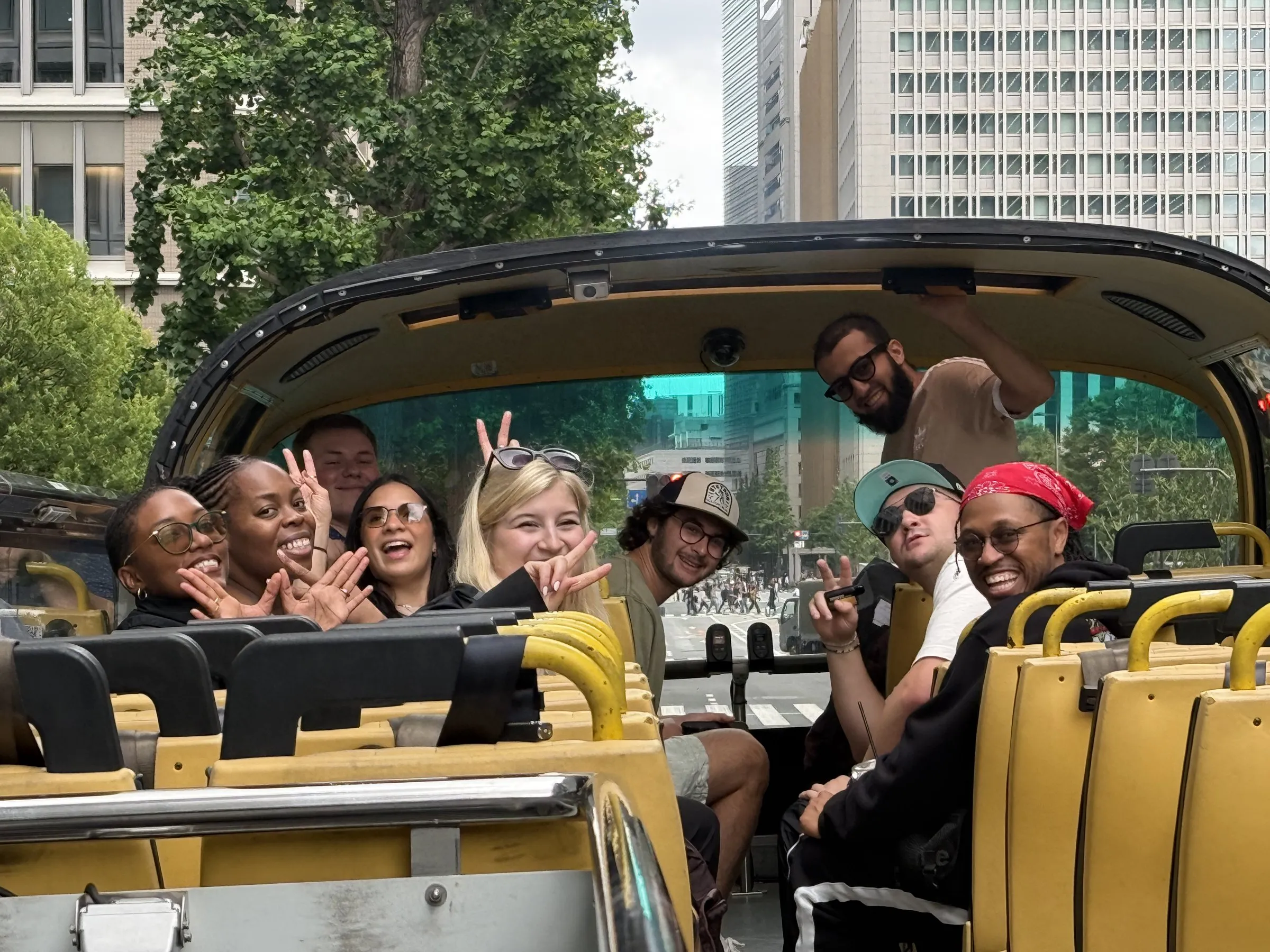 Classmates filling the open upper deck of a Tokyo tour bus, throwing peace signs, with office towers and trees behind them.