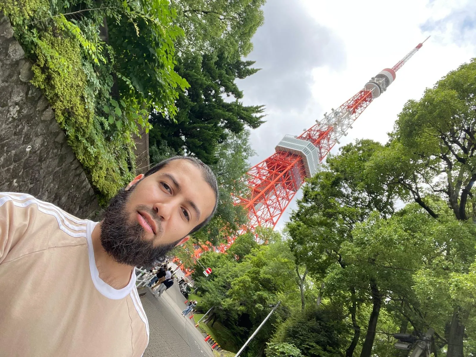 Gotibeh in sunglasses in a park below Tokyo Tower, the orange lattice tower rising behind the trees.