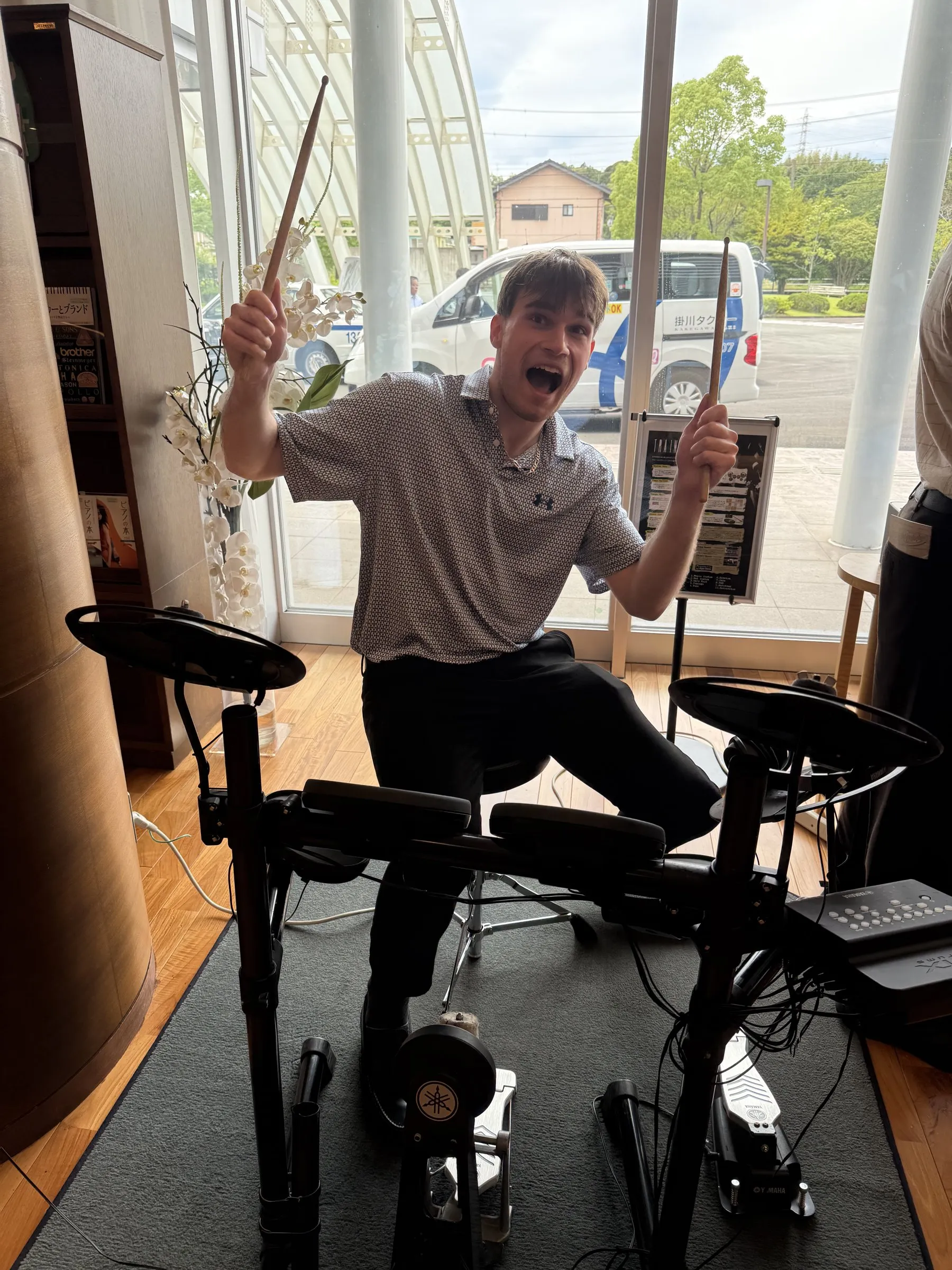 A classmate grinning behind an electronic drum kit in the Yamaha showroom, sticks raised.