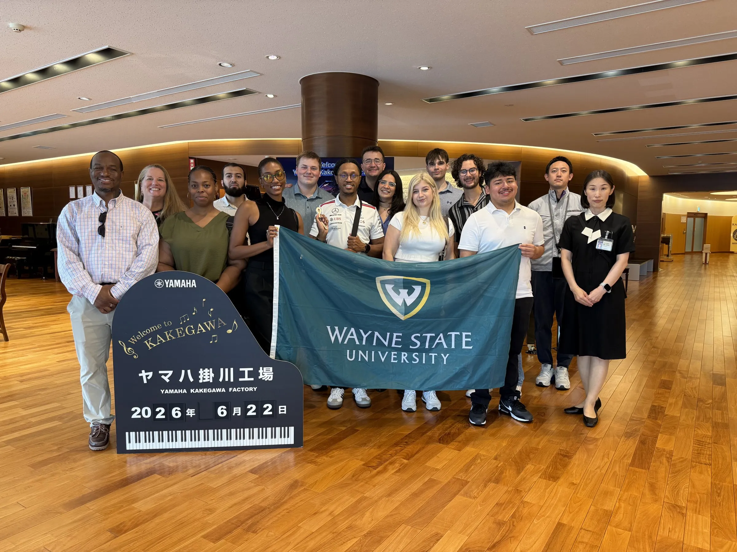 The Wayne State group holding a university flag in the lobby of the Yamaha Kakegawa piano factory, beside a piano-shaped welcome sign dated June 22, 2026.